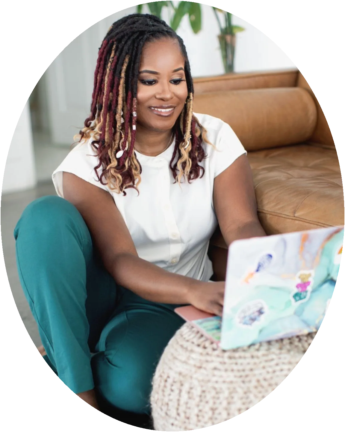 Yvette Howard sitting on the floor beside a tan leather sofa, smiling down at a sticker-covered laptop balanced on a knitted ottoman